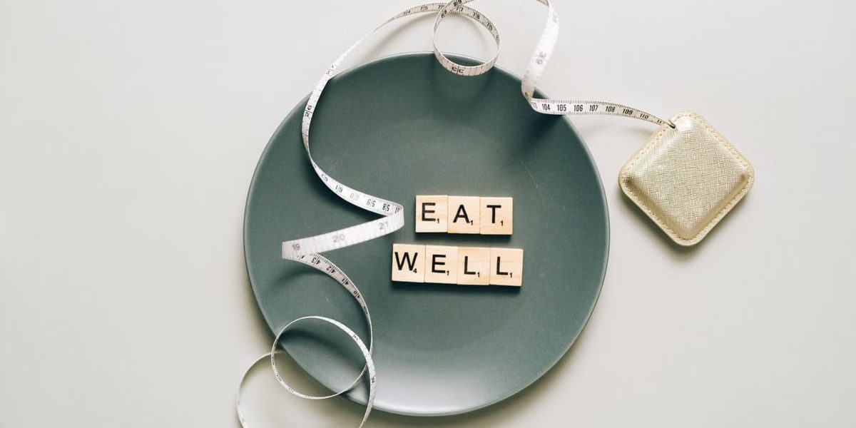 Flat lay of scrabble tiles spelling 'eat well' on a plate with a tape measure, promoting healthy eating.